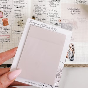 Peut inclure: Un paquet de notes autocollantes transparentes de couleur rose clair, étiquetées "ROSEY BLUSH" et "Transparent Sticky Notes". Les notes sont tenues au-dessus d'un agenda ouvert avec des notes manuscrites et des autocollants décoratifs.