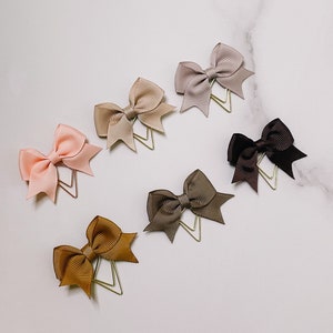 May include: Six different colored bows with gold clips. The bows are made of satin ribbon and are arranged in a row on a white marble surface.