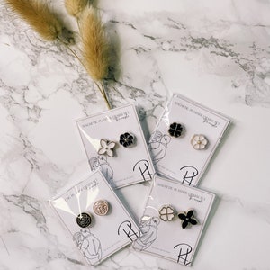 May include: Four sets of magnetic planner charms featuring different flower designs. Each set includes three charms in black, white, and pink. The charms are packaged on white cards with the text "Magnetic Planner Charm Set" and a logo.