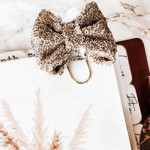 Könnte beinhalten: Ein goldfarbener Büroklammer mit einer glitzernden goldenen Schleife daran. Die Schleife besteht aus Pailletten und liegt auf einer weißen Oberfläche mit einem Blumendesign.