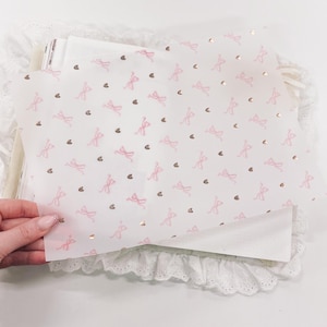 May include: A sheet of white paper with a repeating pattern of pink bows and gold hearts. The paper is slightly translucent and has a delicate, feminine feel.