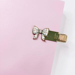 May include: A pink notebook with a decorative bow clasp. The bow is white with a plaid pattern and green trim. The clasp is gold-colored and has a ring for closure.
