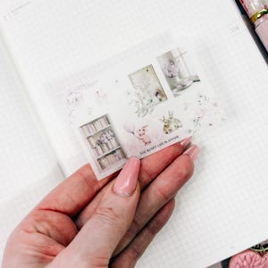 Op de afbeelding: Een decoratieve sticker vellen met aquarel illustraties van konijntjes, boekenplanken en bloemstukken. De tekst "THE ROSEY LIFE PLANNER" is op de sticker vellen gedrukt. Het kleurenpalet omvat zachte roze, witte en groene tinten.