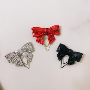 Könnte beinhalten: Drei dekorative Büroklammern in Form von Schleifen. Die Schleifen sind silber, rot und schwarz.