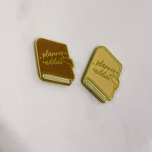 May include: Two gold-coloured enamel pins shaped like closed books. Both pins have the text "planner+addict" in a cursive font. One pin is brown and the other is beige.