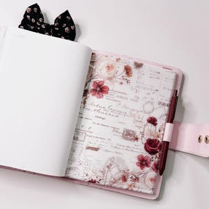Könnte beinhalten: Offener rosa Planer mit floralem Design und Stift. Der Planer hat einen gemusterten Einband mit Rosen und Vintage-Schrift. Eine schwarze Schleife mit floralem Muster ist oben am Planer befestigt.