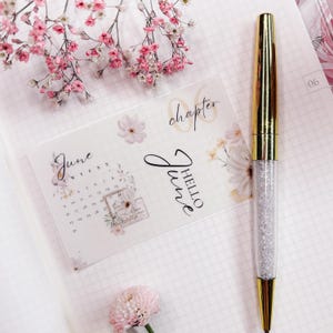 Könnte beinhalten: Ein goldener und kristallener Stift liegt auf weißem kariertem Papier neben einer floralen Kalenderkarte mit der Aufschrift "Juni". Rosa Blumen und Zweige sind um den Stift und die Karte angeordnet. Auf der Karte steht auch "Kapitel" und "Hallo Juni".