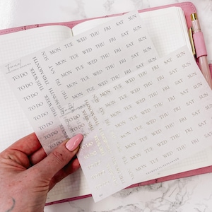Op de afbeelding: Een vel met transparante planner stickers met gouden letters. De stickers bevatten de dagen van de week, "To Do" en "Deze Week".