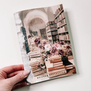 Puede incluir: Un cuaderno de colección de pegatinas con una ilustración de acuarela de una biblioteca con libros y arreglos florales. El cuaderno tiene las palabras "STICKER COLLECTION" impresas en la parte inferior.