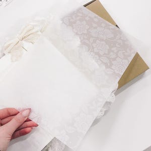 Puede incluir: Papel de seda blanco con un patrón floral, doblado sobre una servilleta de encaje blanco. El papel de seda es translúcido y el patrón floral es visible a través del papel.