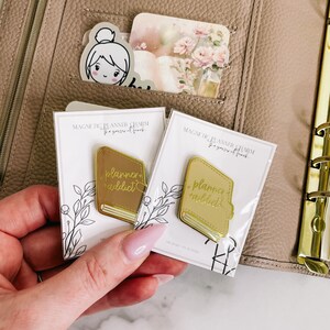 May include: Two gold planner charms with a book design and the text "planner addict" on a white background. The charms are magnetic and are designed to be used in a planner.