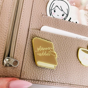 May include: A gold-coloured enamel pin shaped like a closed book with the text "planner+addict" in white script. The pin is attached to a brown leather planner.