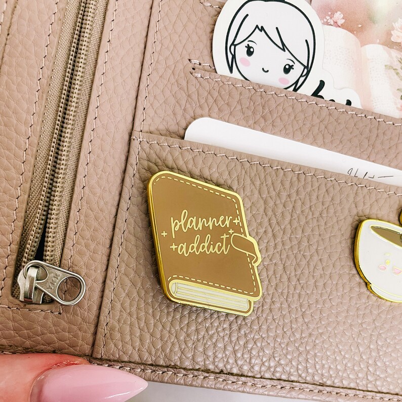 May include: A gold-coloured enamel pin shaped like a closed book with the text "planner+addict" in white script. The pin is attached to a brown leather planner.