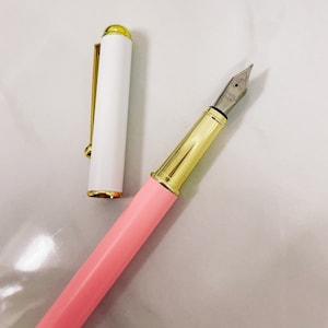 Op de afbeelding: Een roze en gouden vulpen met een witte dop en een gouden penpunt. De penpunt is gemarkeerd met "Stainless Steel Nib".