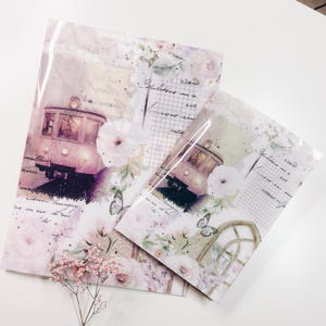 Puede incluir: Dos hojas de papel decorativas con un diseño vintage de tren, flores y mariposas. Las hojas presentan una paleta de colores suaves de rosa, blanco y lavanda, con patrones de escritura y cuadrícula. La hoja más grande mide aproximadamente 21,6 x 27,9 cm.
