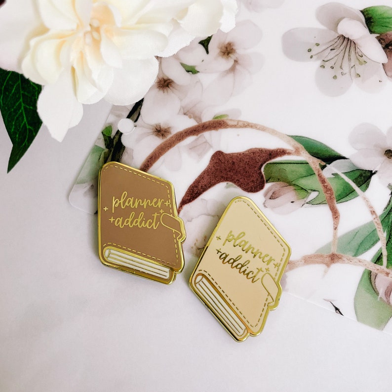 May include: Two gold-toned enamel pins shaped like closed books. Both pins have a brown background and gold lettering that reads "planner + addict".
