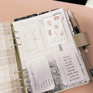 Pu&ograve; includere: Un'agenda rosa e bianca con un tracker di abitudini in bianco e nero, un adesivo di citazione in bianco e nero che dice "Sei il CEO della tua vita. Alzati, bevi un caff&egrave; e fallo." e un disegno a linea in bianco e nero di una persona. L'agenda ha un raccoglitore ad anelli dorato e un'etichetta nera che dice "BIG BOSS".