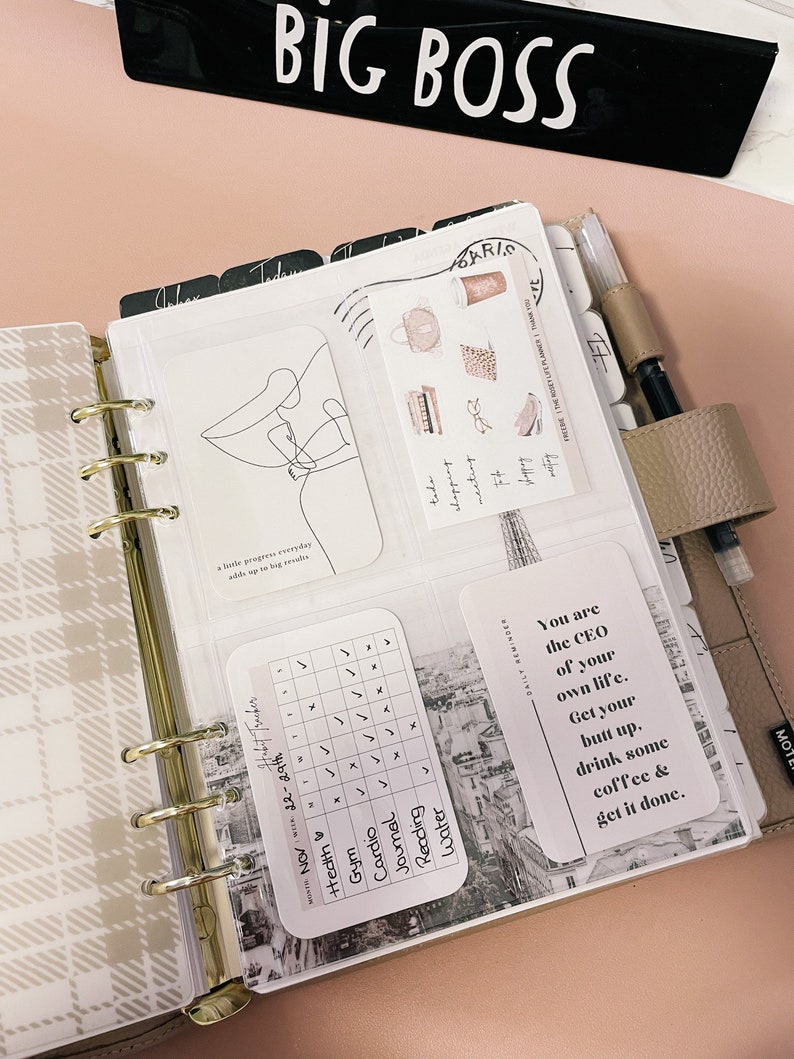 Pu&ograve; includere: Un'agenda rosa e bianca con un tracker di abitudini in bianco e nero, un adesivo di citazione in bianco e nero che dice "Sei il CEO della tua vita. Alzati, bevi un caff&egrave; e fallo." e un disegno a linea in bianco e nero di una persona. L'agenda ha un raccoglitore ad anelli dorato e un'etichetta nera che dice "BIG BOSS".