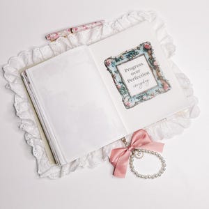 May include: An open journal with a white lace cover and a pink ribbon bookmark. The journal page has a decorative frame with the text "Progress over Perfection everyday".