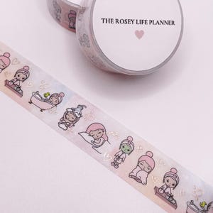 May include: A pink and white washi tape with illustrations of people in various activities, including taking a bath, sleeping, and applying a face mask. The tape also features gold foil accents and the text "THE ROSEY LIFE PLANNER".