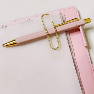 Può includere: Un'agenda in pelle rosa con divisori dorati e una penna rosa con un fermaglio dorato. I divisori sono etichettati "Obiettivi", "Routine", "Università" e "Fatture".