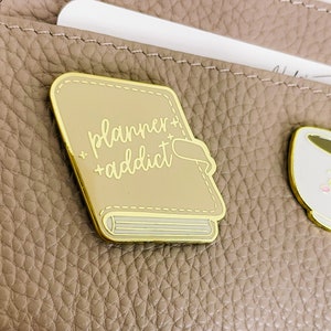 May include: Gold-coloured enamel pin shaped like a closed book with the text "planner + addict" in white script on the cover.