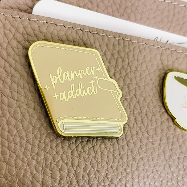 May include: Gold-coloured enamel pin shaped like a closed book with the text "planner + addict" in white script on the cover.