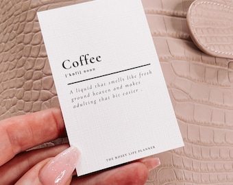 May include: A white card with the word "Coffee" written in black text. The card also includes a definition of coffee as a liquid that smells like fresh ground heaven and makes adulting a bit easier. The card is on a pink textured surface and the text "THE ROSEY LIFE PLANNER" is at the bottom of the card.