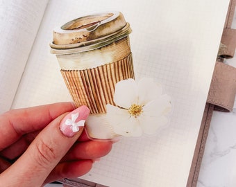 May include: A clear sticker with a brown and white coffee cup and a white flower. The coffee cup has a brown lid and a brown and white striped design. The flower has five petals and a yellow center.