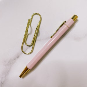 Pu&ograve; includere: Una penna rosa con un cappuccio dorato e una graffetta dorata su una superficie di marmo bianco.