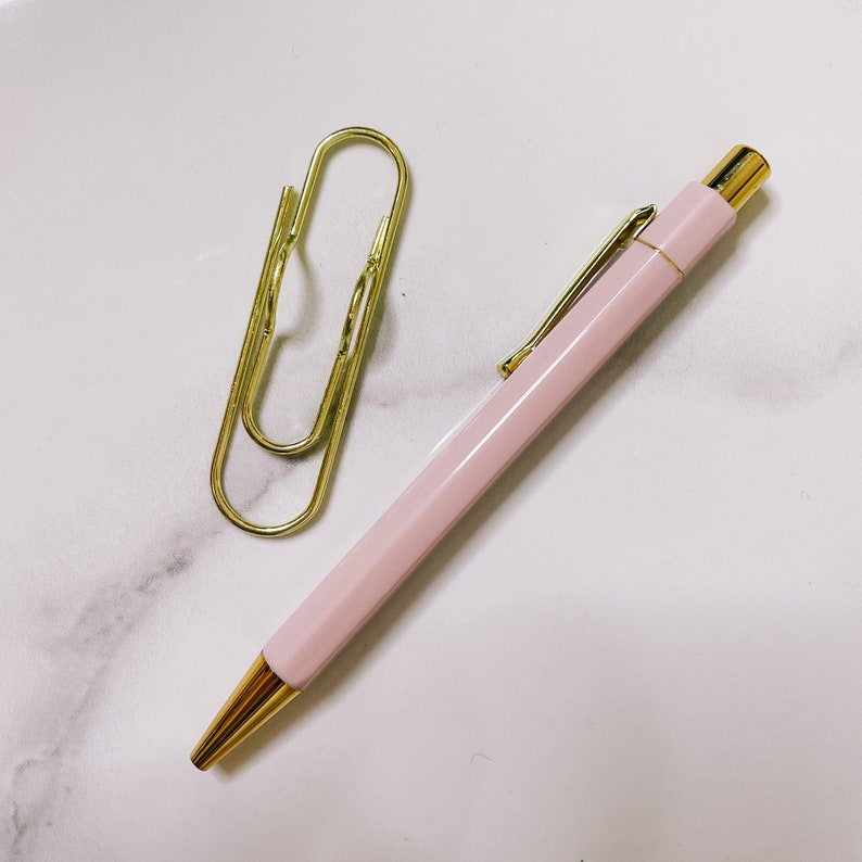 Pu&ograve; includere: Una penna rosa con un cappuccio dorato e una graffetta dorata su una superficie di marmo bianco.