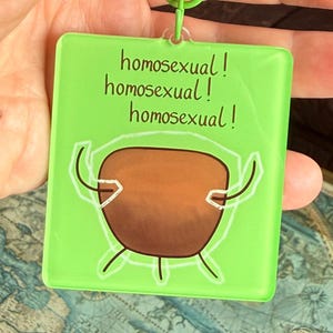 May include: A square lime green acrylic keyring with a lobster clasp. The keyring features a brown, rounded, cartoon-like shape with thin white outlines and the text "homosexual! homosexual! homosexual!" printed in a dark brown, handwritten font.
