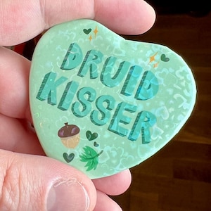 May include: A green heart-shaped button with the text "Druid Kisser" in a blue font. The button has a white speckled background with small hearts and acorns.