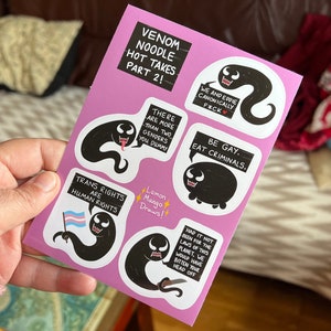 Pode incluir: Uma folha de papel rosa com vários adesivos em preto e branco que apresentam um personagem de desenho animado com um balão de fala. Os balões de fala contêm texto como "Venom Noodle Hot Takes Part 2!" e "Trans Rights Are Human Rights".