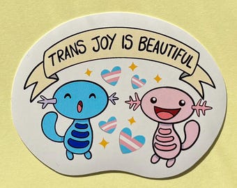 Puede incluir: Una pegatina con dos ajolotes de dibujos animados, uno azul y otro rosa, con el texto "TRANS JOY IS BEAUTIFUL" en una pancarta. El diseño incluye corazones con los colores de la bandera transgénero y estrellas.