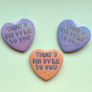 以下が含まれることがあります： テキスト入りのハート型のボタンが3つ。2つのボタンは紫で、1つはオレンジ色です。紫色のボタンには「THAT'S MS DYKE TO YOU」と「THAT'S MX DYKE TO YOU」と書かれています。オレンジ色のボタンには「THAT'S MR DYKE TO YOU」と書かれています。