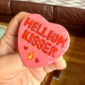 Può includere: Un oggetto rosa a forma di cuore con la scritta "HELLION KISSER" in lettere rosse. Il design include piccoli cuori rossi, stelle gialle e una fiamma. L'oggetto è tenuto in una mano.