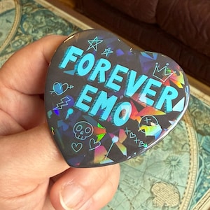 Op de afbeelding: Een hartvormige button met de tekst "FOREVER EMO" in lichtblauw. De donkere achtergrond heeft holografische accenten en kleine, witte doodles van sterren, harten en een schedel.