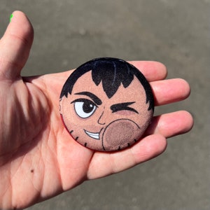 Pode incluir: Um botão redondo com uma ilustração de desenho animado de uma pessoa piscando. A pessoa tem cabelo preto, pele marrom e está vestindo uma camisa vermelha. O fundo do botão é branco.