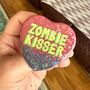 May include: A heart-shaped badge with the words "ZOMBIE KISSER" in green lettering on a red background. The bottom of the badge has a pattern of colourful hearts. The badge is held in a hand.