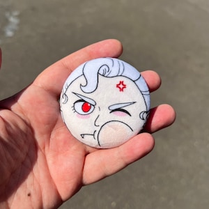 May include: A circular button featuring an angry cartoon face with white hair, a red eye, and a winking eye. The design includes a red angry symbol above the eyebrow. The button is held in a hand.