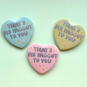 Peut inclure: Trois badges en forme de cœur aux couleurs pastel : bleu clair, jaune et rose. Chaque badge porte l'inscription "THAT'S MS/MX/MR FAGGOT TO YOU" en gris. Des étoiles oranges décorent chaque badge.