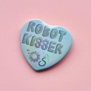 May include: A heart-shaped badge with the text "ROBOT KISSER" in 3D lettering. The badge is light blue with a gradient effect. It features a gear and a stylized "S" with hearts and sparkles. The background is pink.