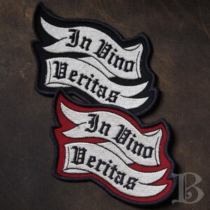 Peut inclure: Deux écussons brodés avec la phrase latine "In Vino Veritas" sur une bannière. L'un des écussons a une bordure noire, l'autre une bordure rouge. Le texte est en police gothique, et la bannière est blanche.