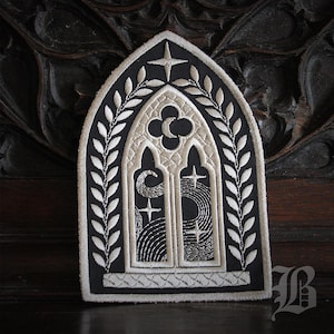 Peut inclure: Écusson brodé représentant une fenêtre gothique en arc. Le patch a un fond noir avec des détails brodés blancs, dont une bordure florale, des étoiles et un croissant de lune. Le patch mesure environ 10 cm de haut.