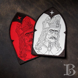 Peut inclure: Deux écussons brodés représentant un homme aux longs cheveux, une moustache et une couronne. L'un est rouge avec une bordure noire, l'autre blanc avec une bordure noire. Les deux ont une étoile en haut.