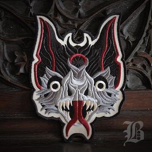 Peut inclure: Écusson brodé représentant une tête de loup stylisée. Le patch a un fond noir avec des détails gris et blancs, des accents rouges et une bordure blanche. La gueule du loup est ouverte, révélant des dents acérées et une langue rouge.