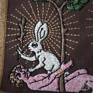 Peut inclure: Écusson brodé représentant un lapin blanc tenant un bâton au-dessus d'une personne. Le motif comprend un arbre et un motif de rayons de soleil, avec du fil marron, doré, blanc, rose et vert sur fond marron. L'écusson a une bordure dorée.