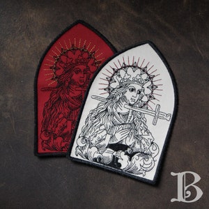 Peut inclure: Deux écussons brodés en forme d'arc gothique. L'un est rouge avec des accents dorés, l'autre est blanc avec des détails noirs et rouges. Ils présentent tous deux une illustration médiévale d'une femme avec une épée et une auréole.