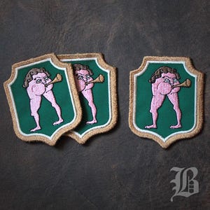 May include: Embroidered patches with a humorous design of a figure playing a horn. The patches are shield-shaped, green with a gold border and white trim. The figure is pink with brown hair.